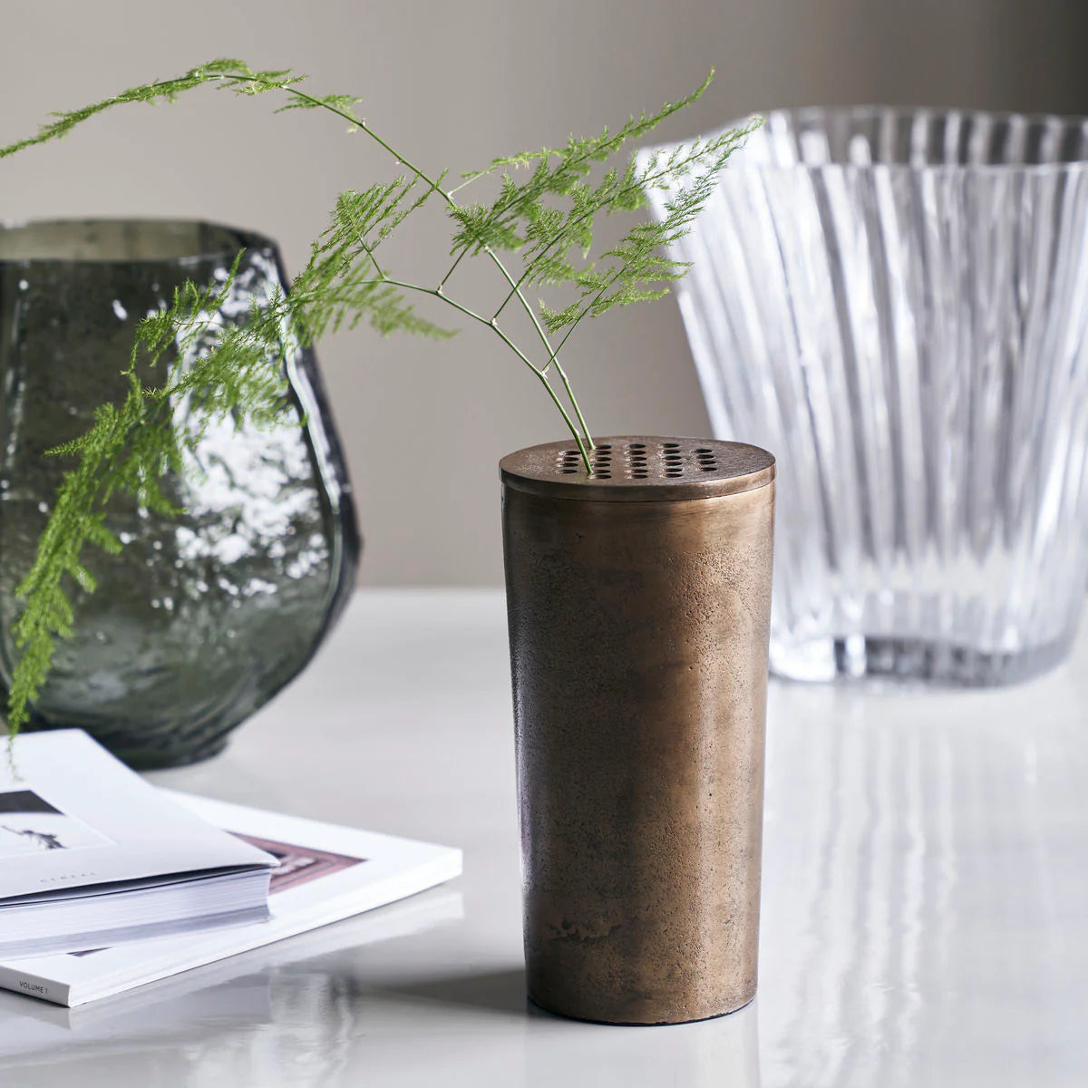 Bronze vase with green plant on a white surface with glass bowls in the background