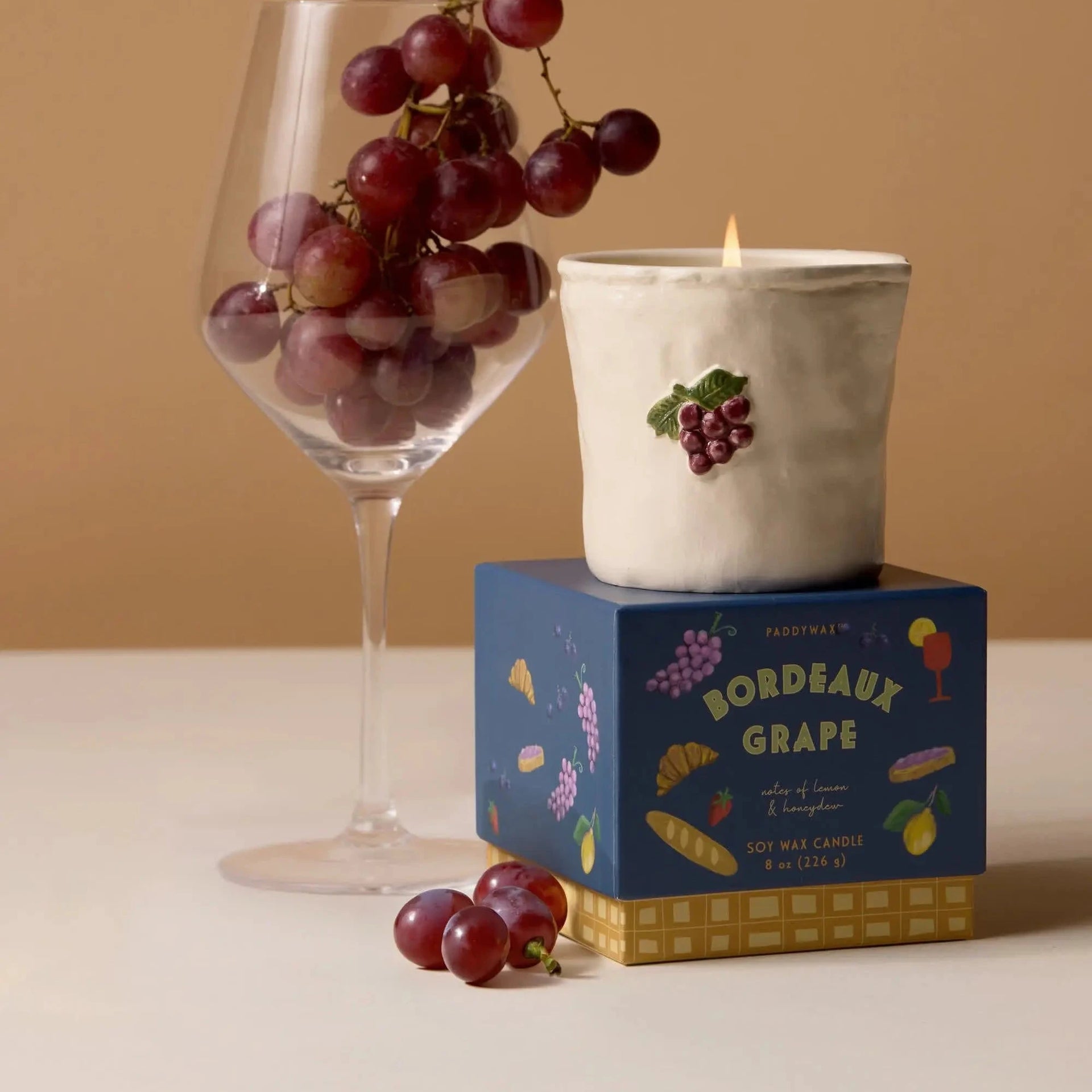 Candle with grape design on a box, surrounded by grapes and a glass against a beige background