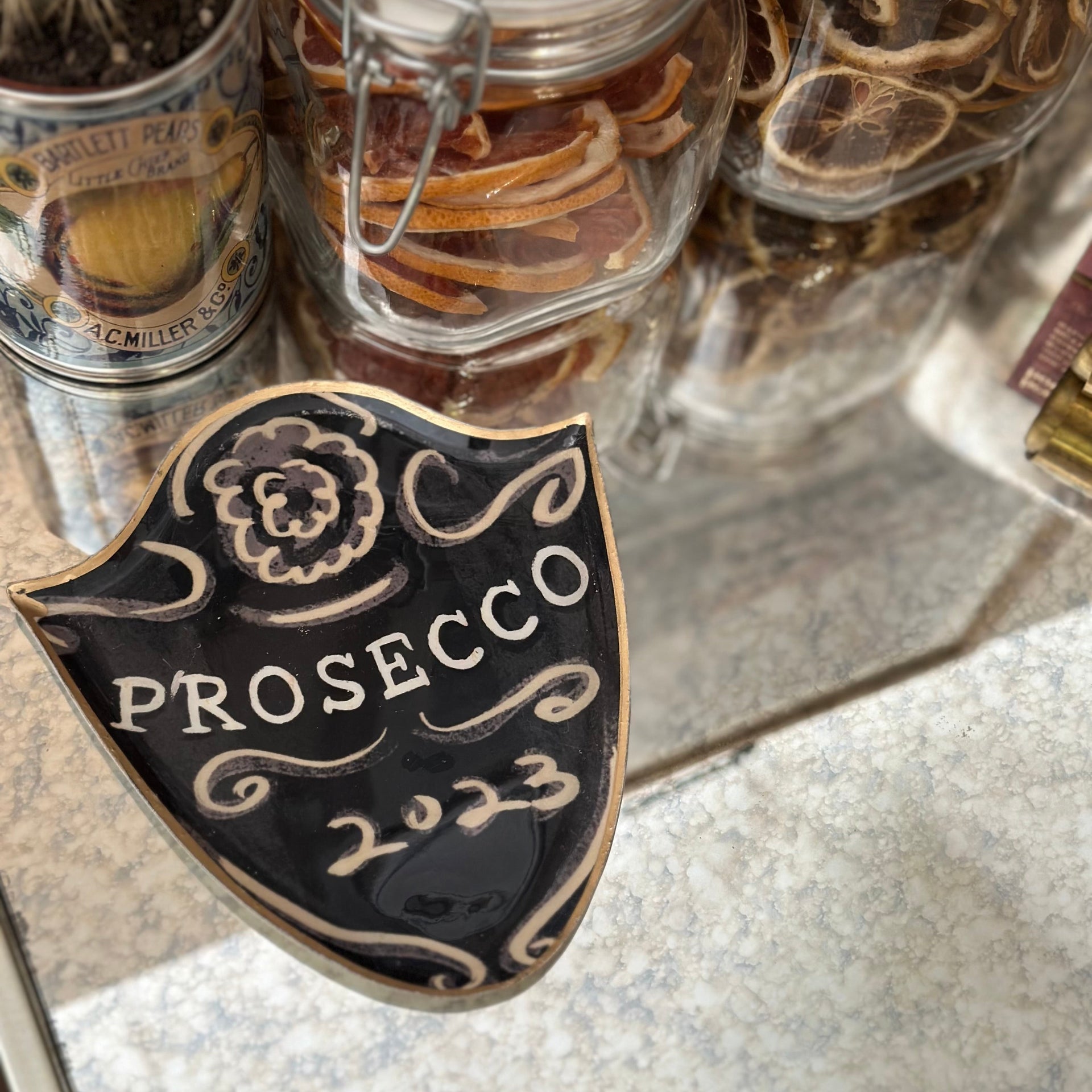 Prosecco 2023 decorative dish on a reflective surface with jars of snacks in the background.